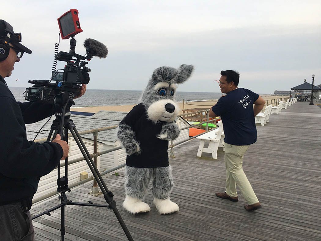 Filming a costumed dog with a camera on a pier; a man gestures beside them. Cloudy sky, ocean.