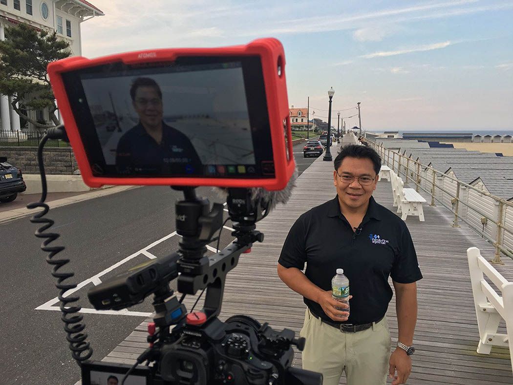 Man reporting on camera at a boardwalk. Holds water bottle, red monitor displays his image.