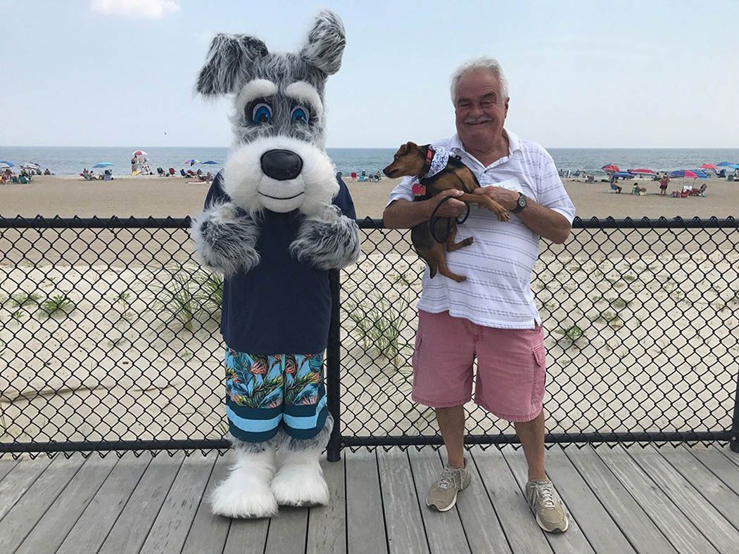 Mascot dog in beach attire stands next to a man holding a small dog on a boardwalk overlooking the beach.