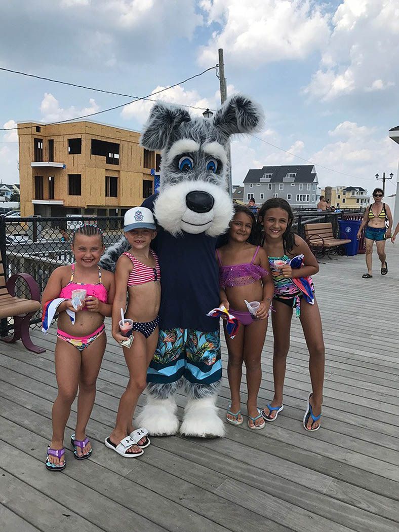 Four girls in swimwear pose with a dog mascot on a boardwalk.