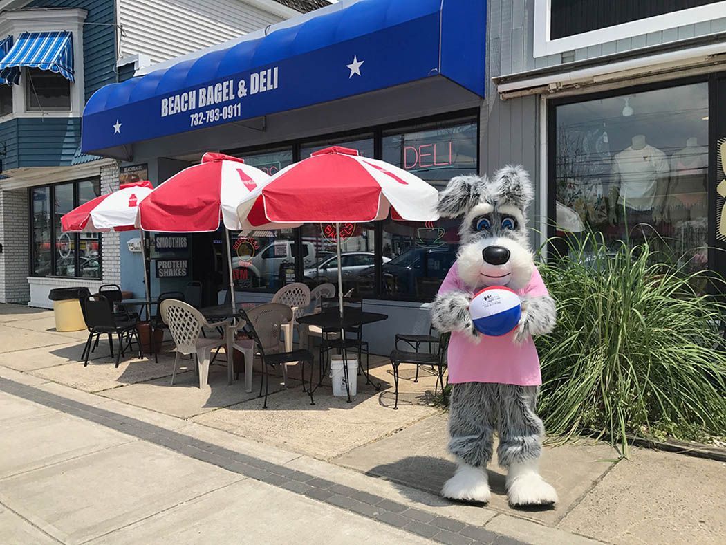 Mascot dog in pink shirt holds beach ball in front of restaurant with outdoor seating.