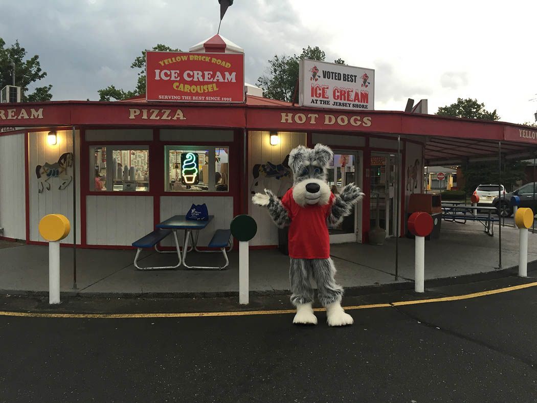 Mascot dog waves in front of a red ice cream stand with signs for ice cream, pizza, and hot dogs.