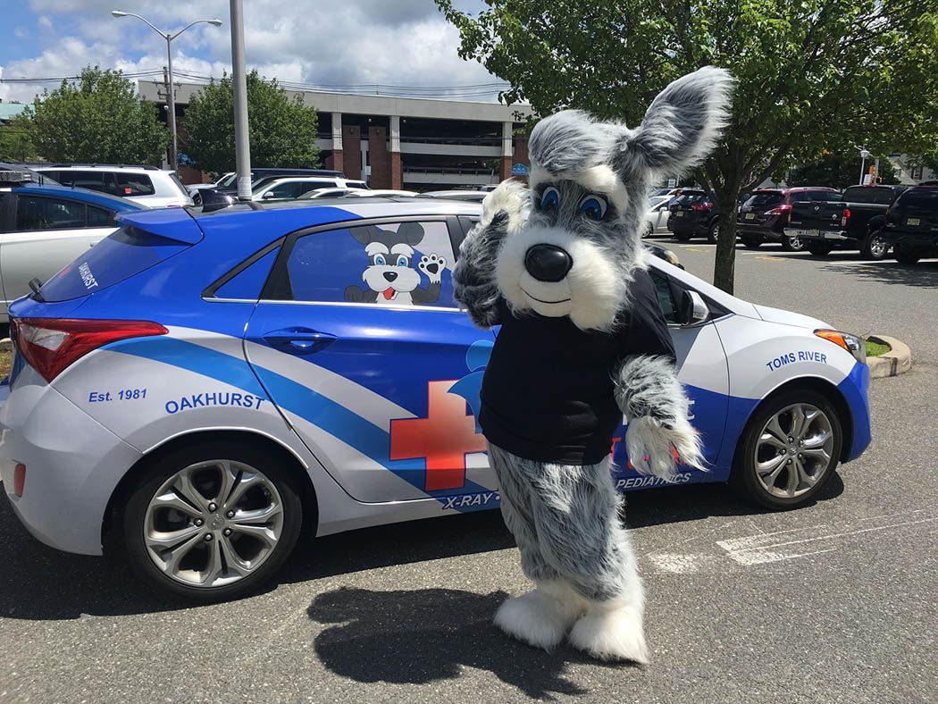 A mascot in a dog costume poses beside a blue and white car with dog graphics.
