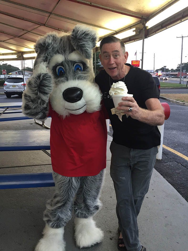 Man smiling with soft-serve ice cream, posing with a dog mascot outside a restaurant.