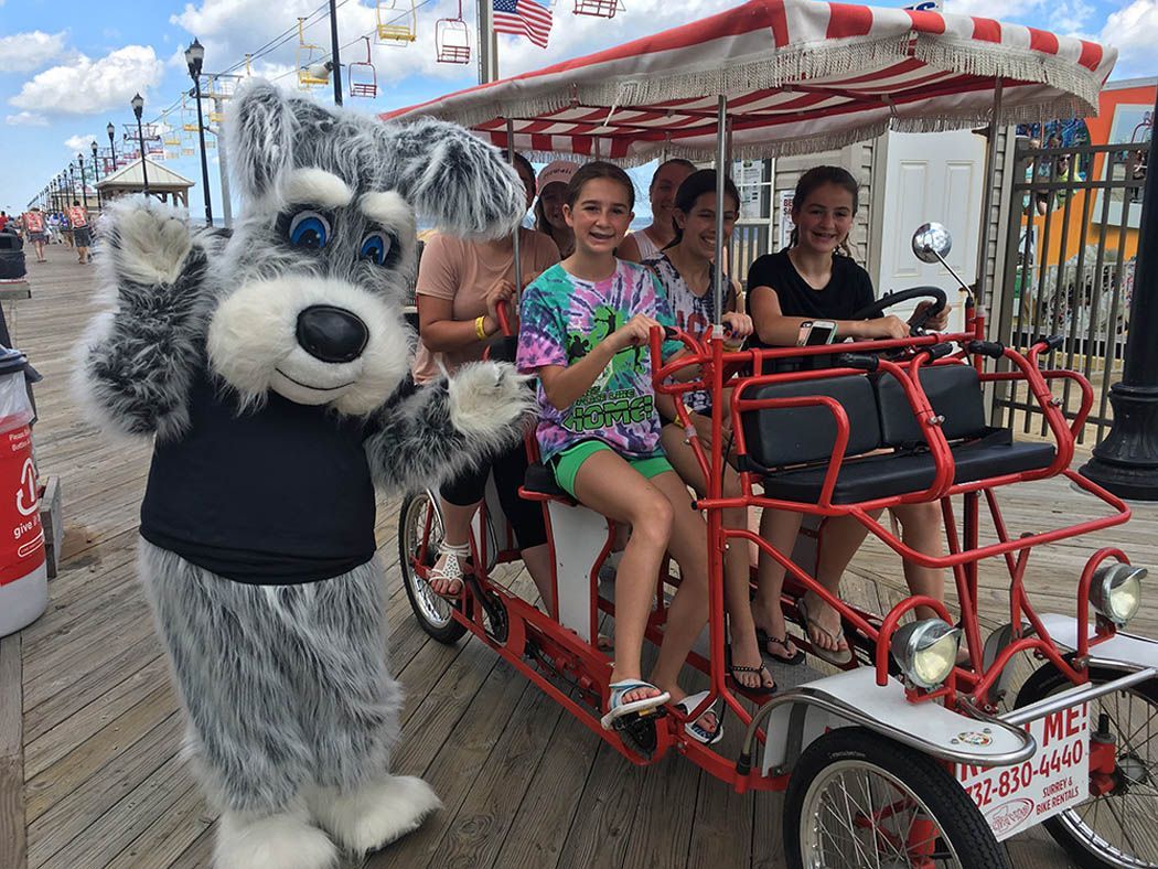 Mascot waves at a surrey bike with children on a boardwalk.