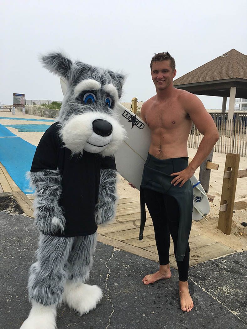 A man with a surfboard poses next to a husky mascot on a beach.