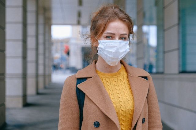 Woman wearing a face mask and tan coat stands outside a building.