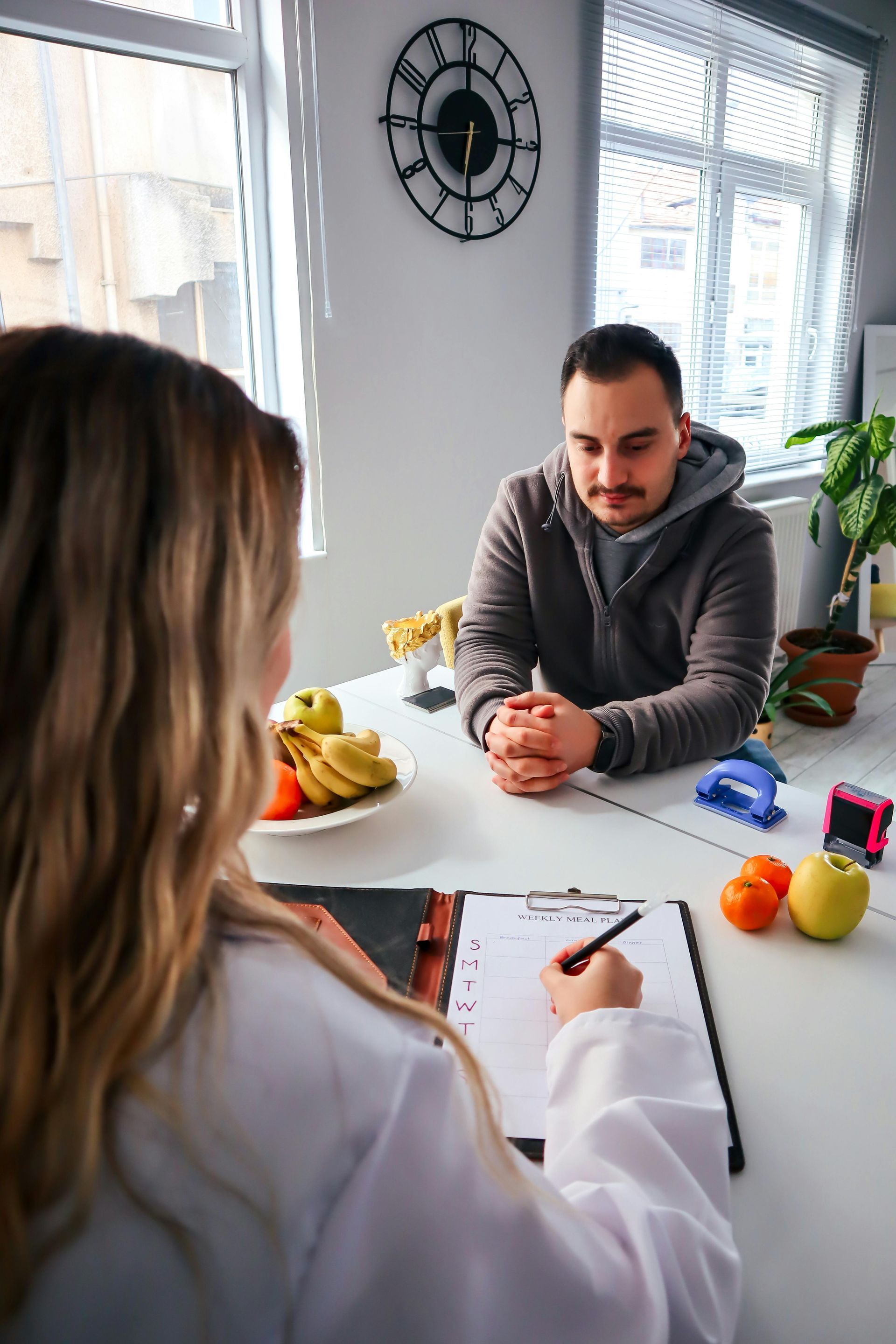 Two people talking across a desk in a bright office, with fruit, notes, and a clock on the wall.