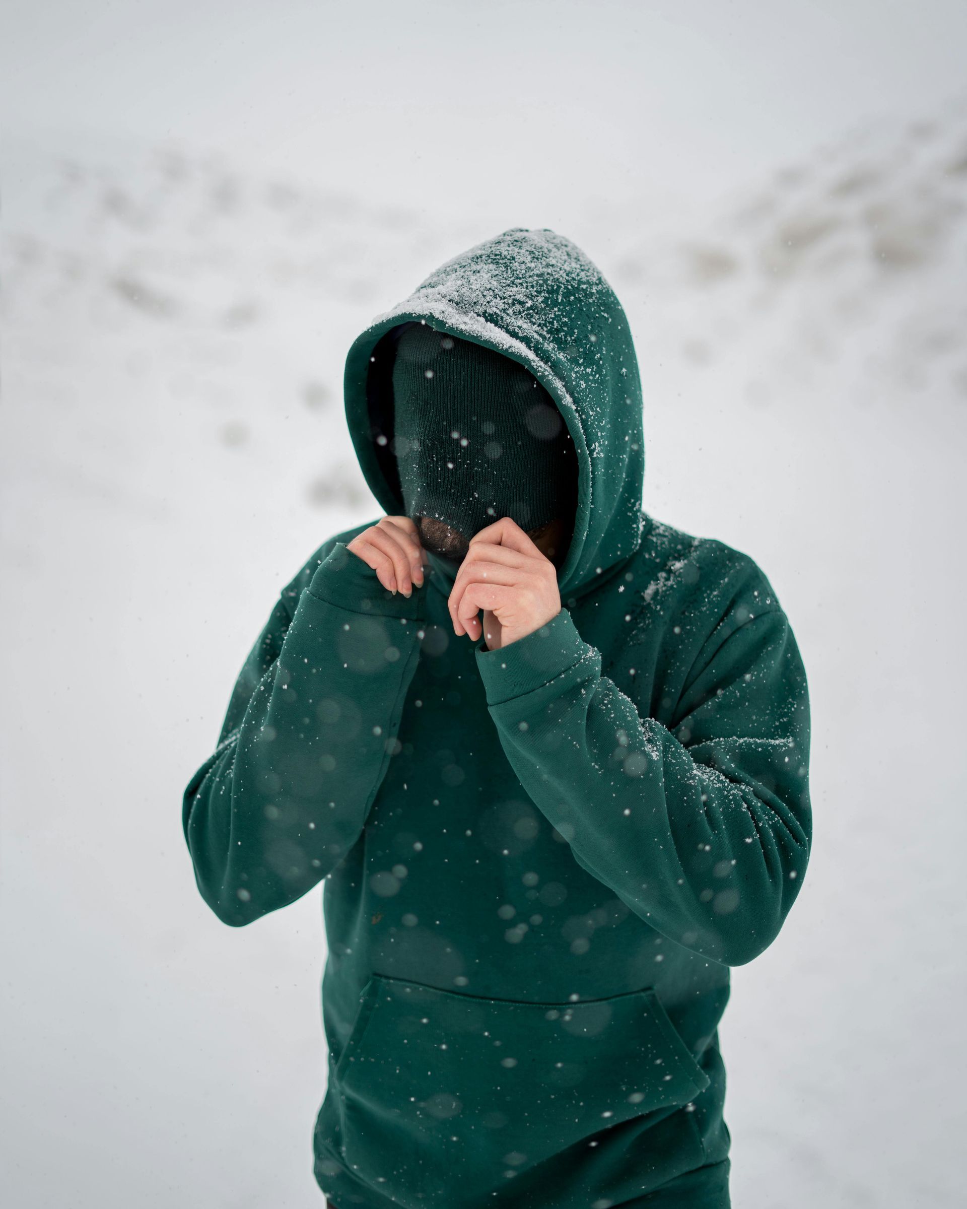 Person in a green hoodie pulling the hood tight in falling snow.