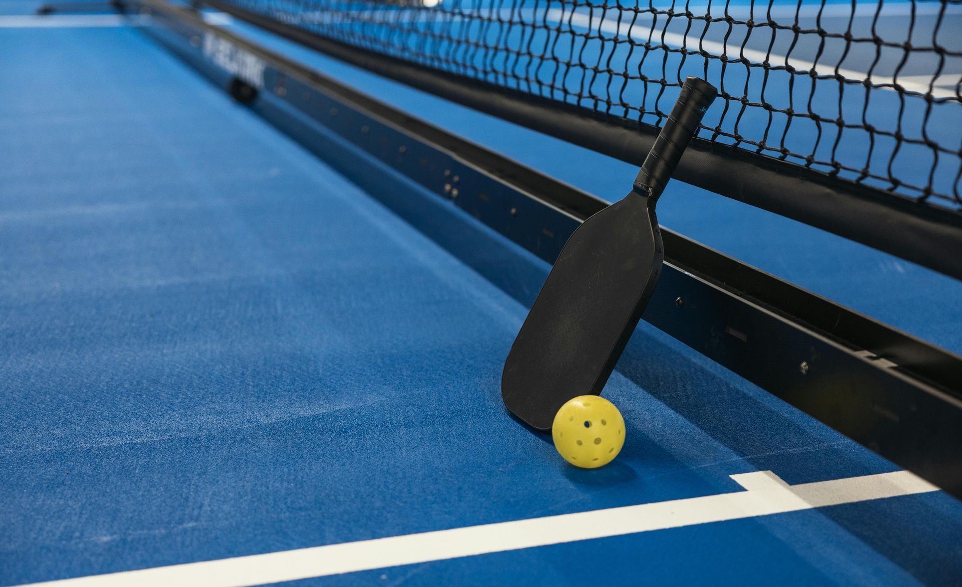 Pickleball paddle leaning against net beside a yellow ball on a blue court