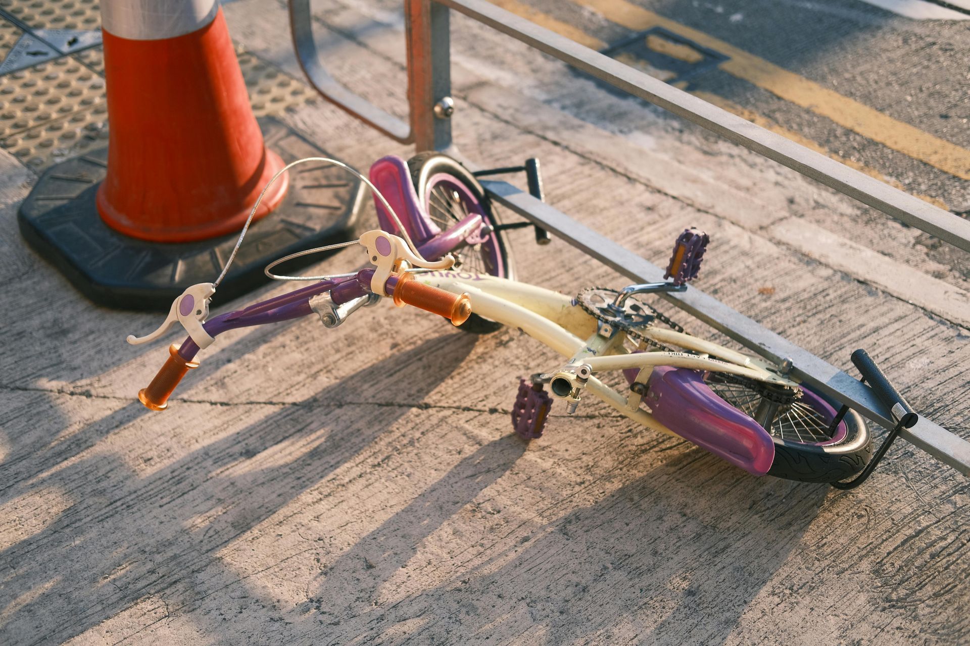 Broken pink-and-purple bicycle lying on a sunlit sidewalk beside an orange traffic cone