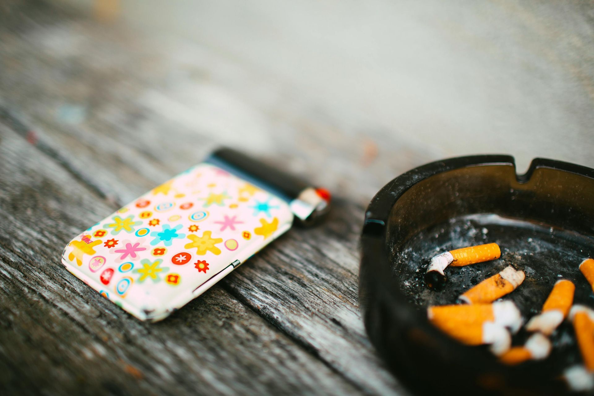 Floral-patterned cigarette case beside an ashtray with cigarette butts on a wooden table.