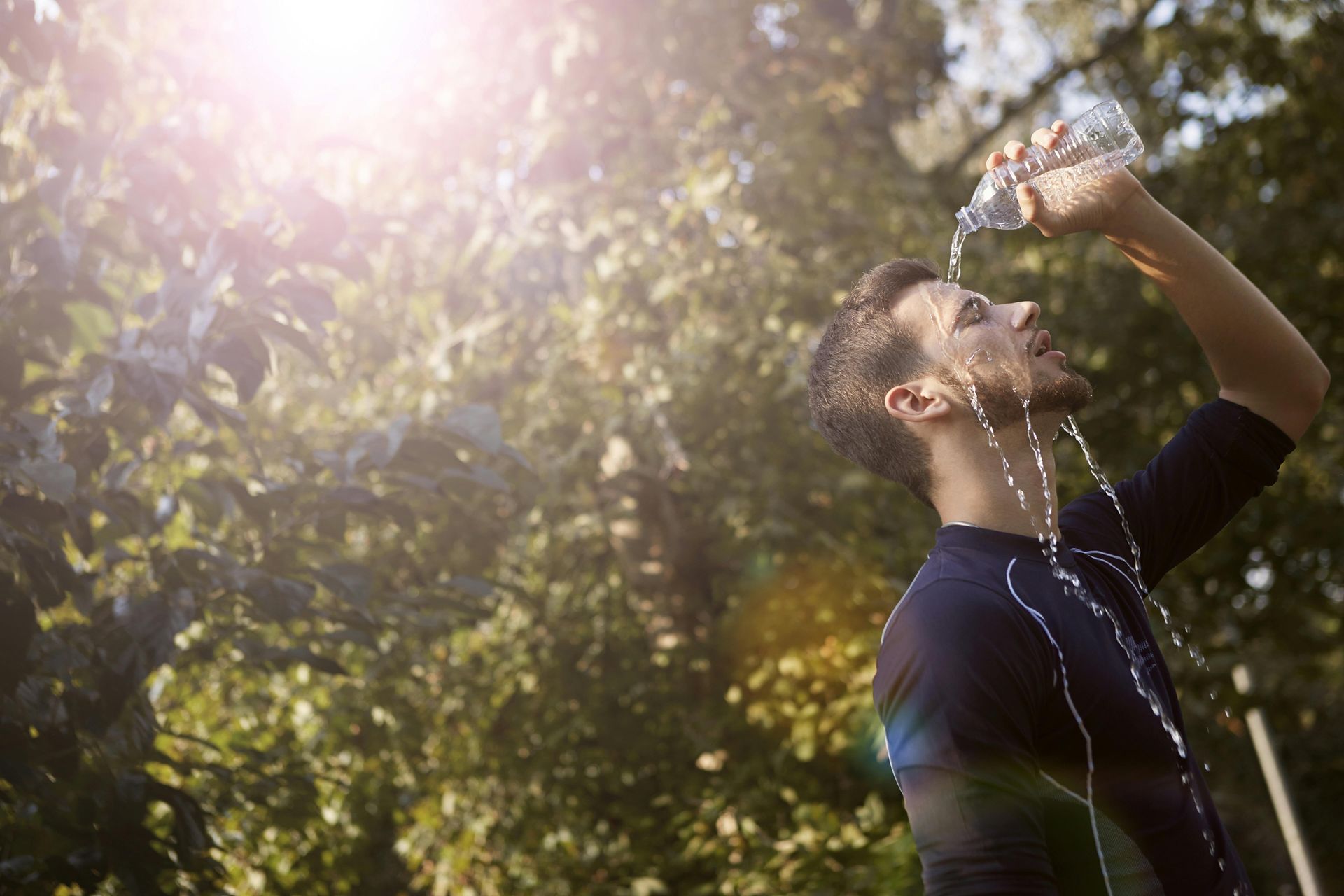 Person drinking water outdoors in sunlight, with trees and bright lens flare in the background