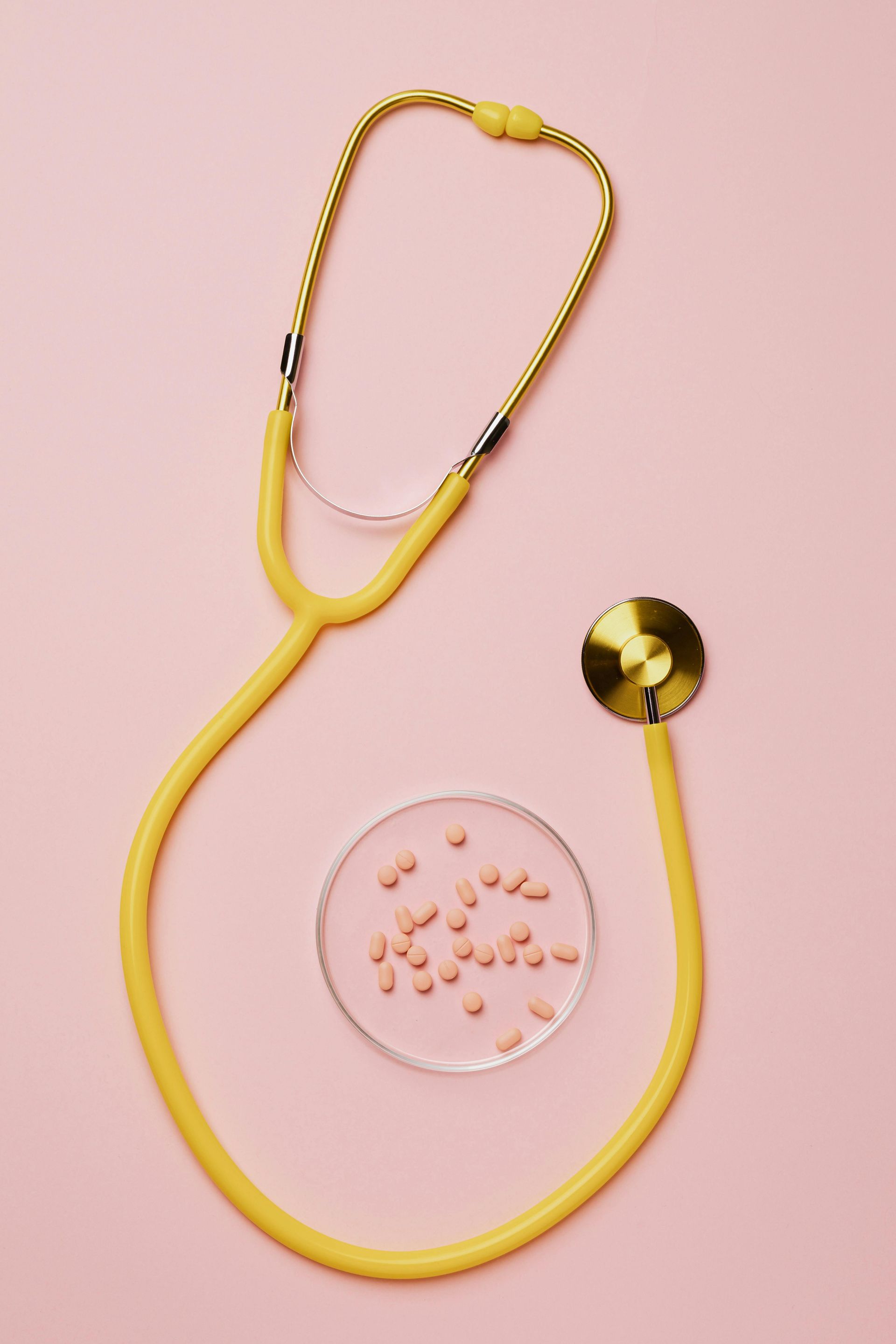 Yellow stethoscope with a gold chestpiece on a pink background beside a petri dish of pills.