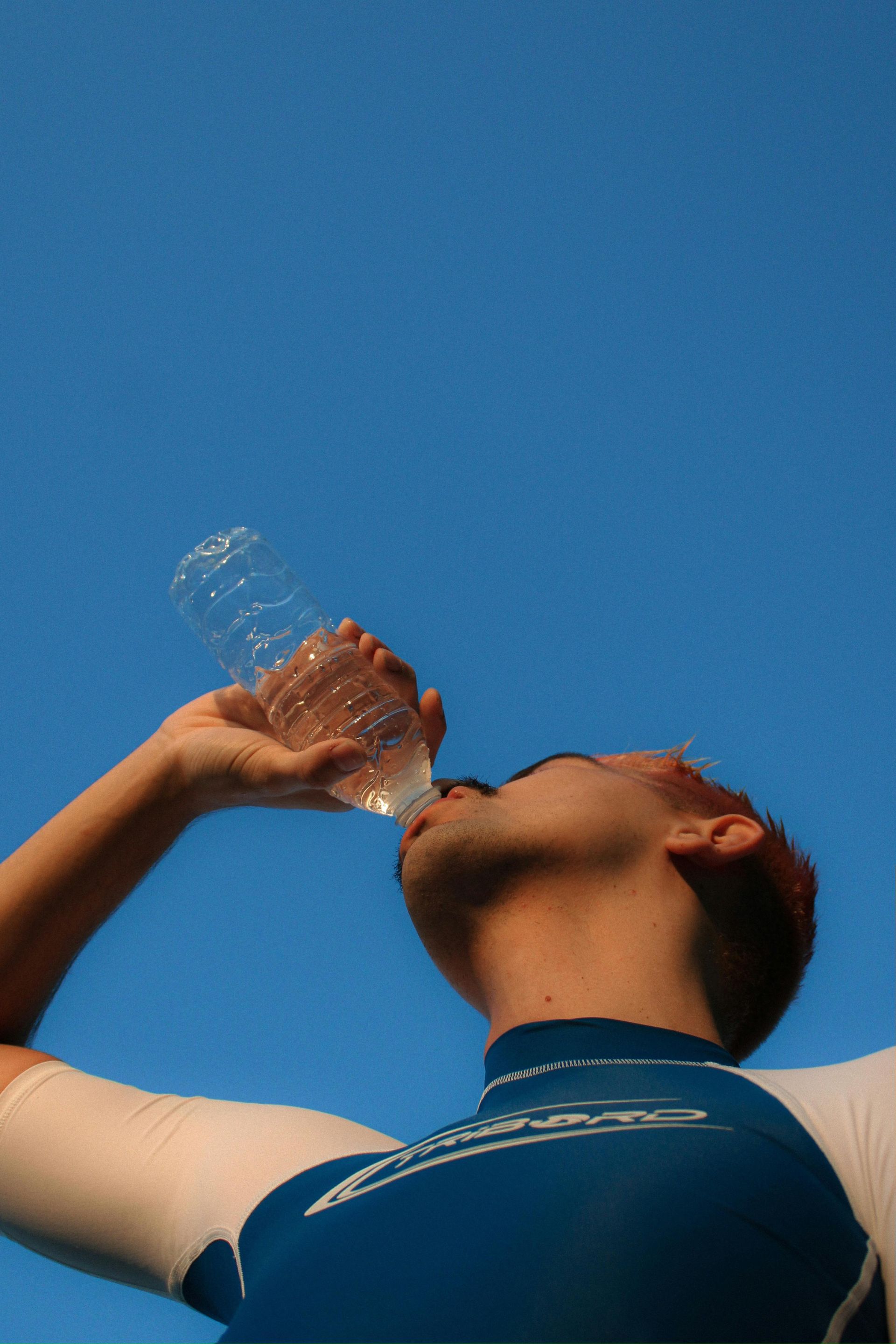 Person drinking from a water bottle against a clear blue sky.