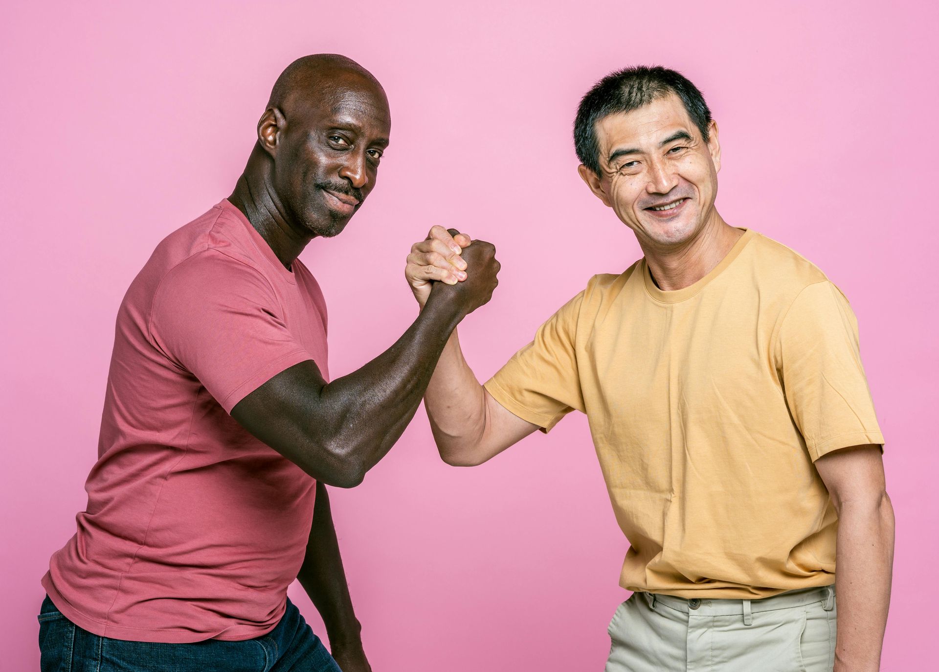 Two people arm wrestling against a pink background, smiling and leaning in.