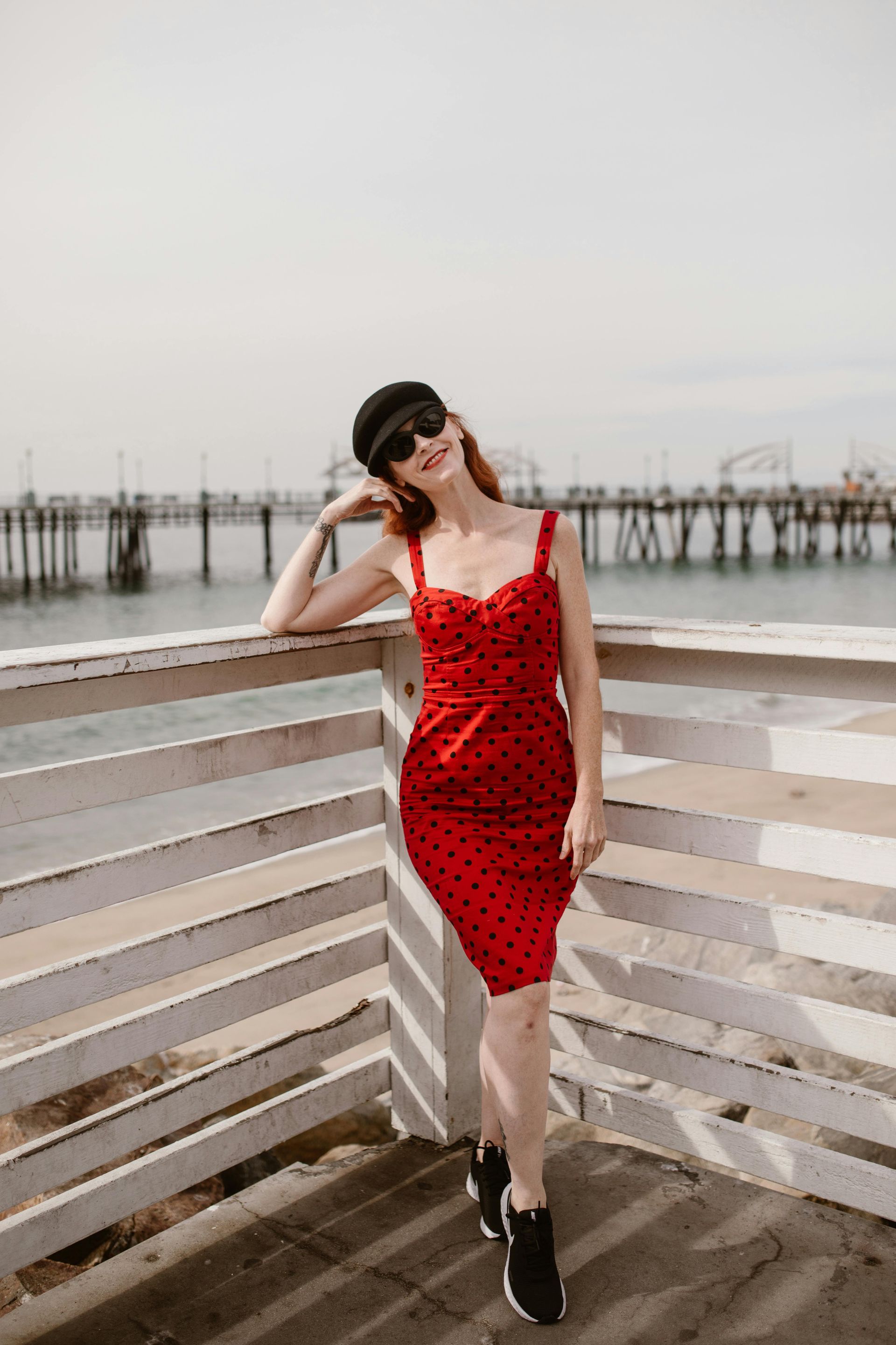 Person in a red dress and sunglasses posing on a waterfront boardwalk near the pier.
