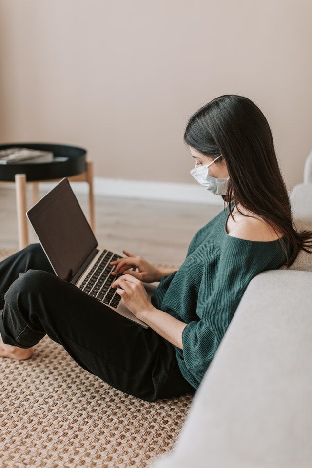 Woman wearing face mask works on laptop while seated on floor near a couch.
