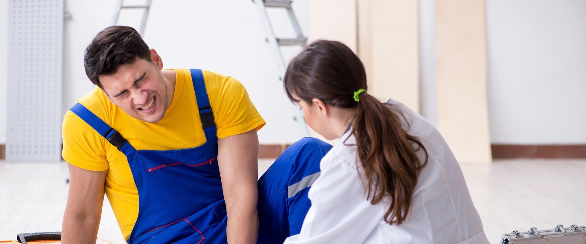 A man in work clothes grimaces in pain as a woman in a lab coat examines his leg. Indoors, near a ladder.
