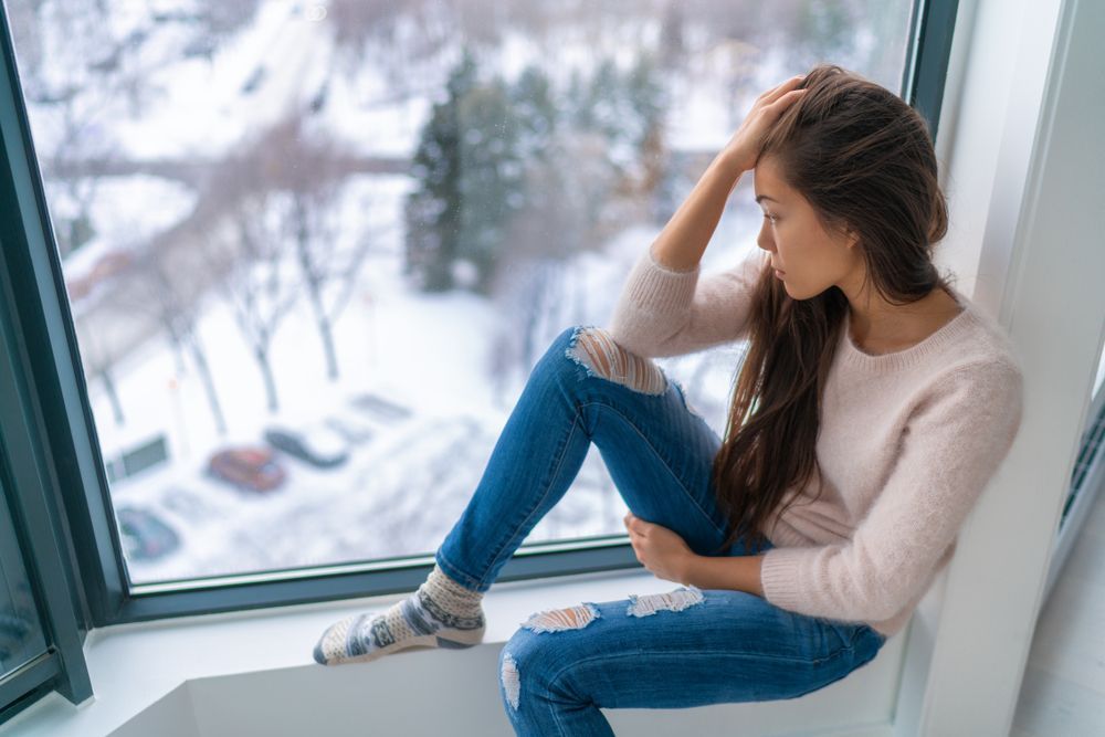 Woman sitting in a window, head in hand, looking out at a snowy landscape, appearing pensive.
