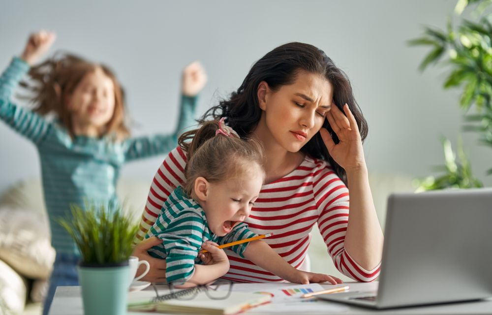 Woman looking stressed with two children. One child is crying and the other is raising their arms. Laptop on desk.