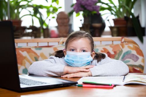 Girl wearing a mask, looks at the camera, leaning on desk with laptop and notebook.