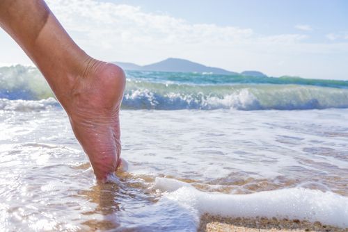 Foot entering ocean water on a sunny beach, waves and distant mountains in the background.