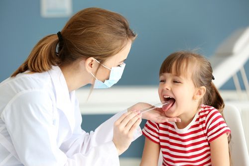 Doctor examining a child's throat with a tongue depressor in a medical office setting.