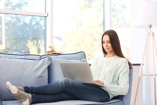 Woman on a couch with a laptop, typing, in a well-lit living room.