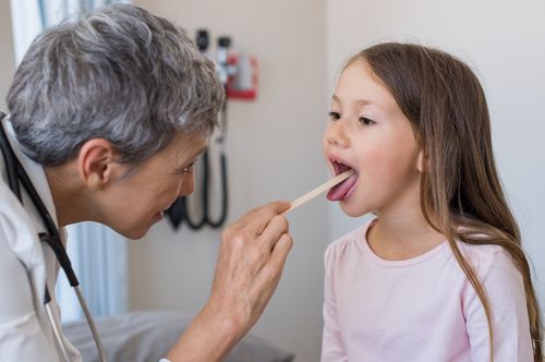 Doctor examining a child's throat with a tongue depressor in a medical examination room.