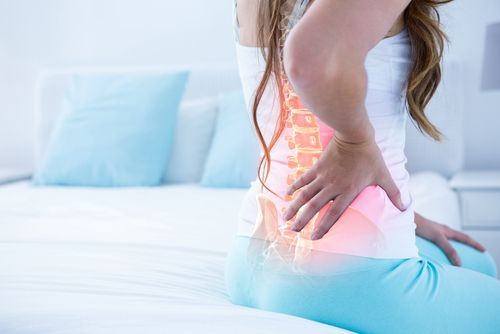 Woman sitting on a bed, clutching her lower back in pain, highlighted spine and pelvis.