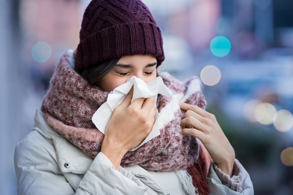 Woman blowing her nose with a tissue outside in cold weather, wearing a hat, scarf, and coat.