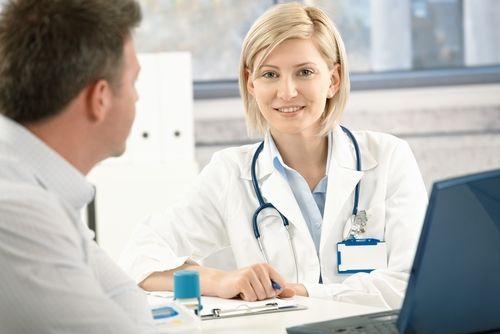 Doctor and patient in an office setting; doctor in white coat, smiling, laptop on desk.
