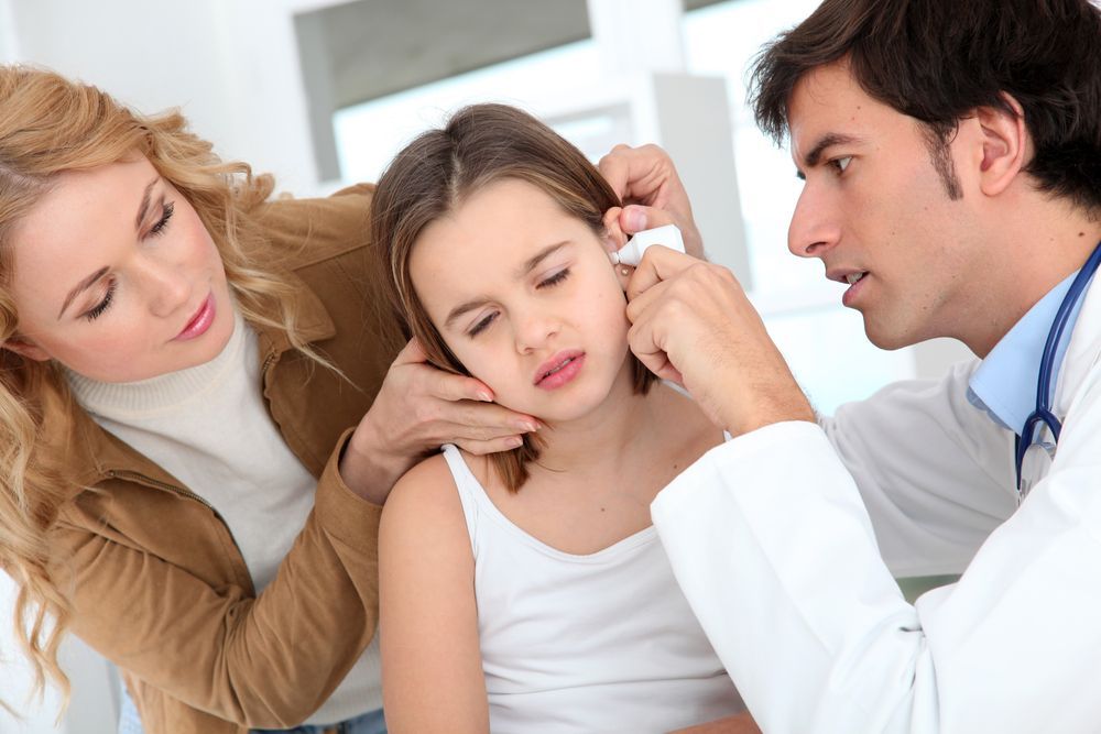 Doctor examining a girl's ear, mother watches. Girl appears to be in discomfort, medical setting.