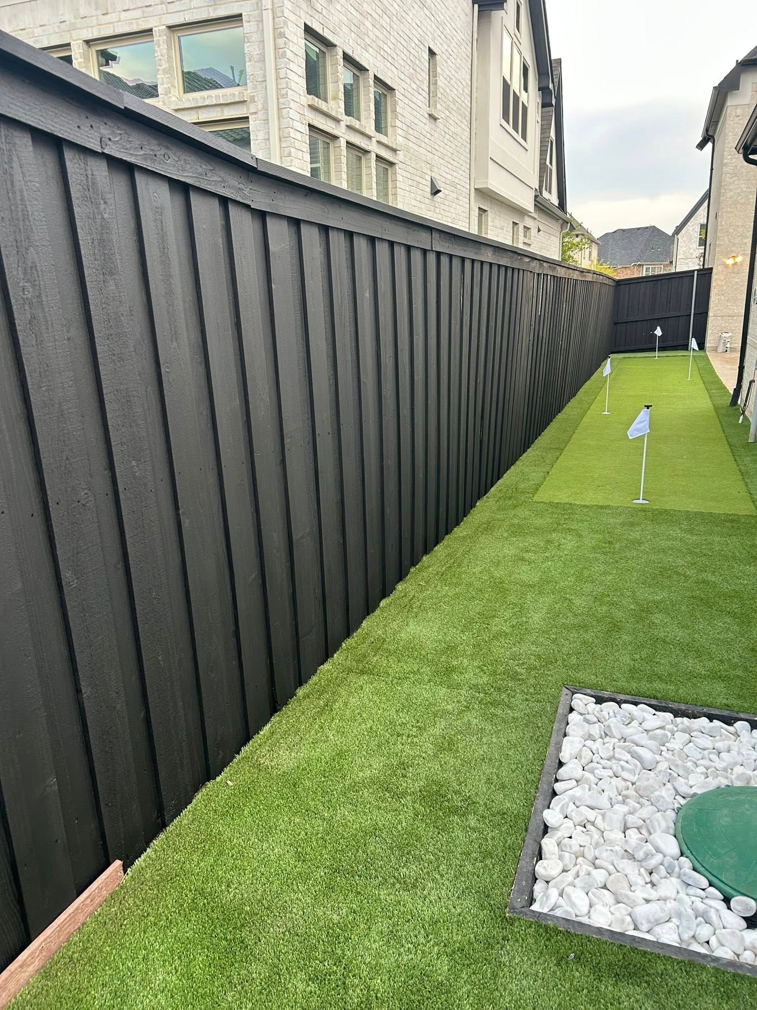Narrow artificial grass side yard with black fence, apartment buildings, and a small gravel feature.