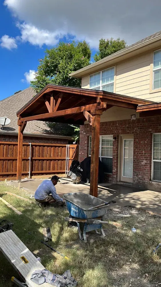 Person working on a wooden patio cover in a backyard next to a two-story house.