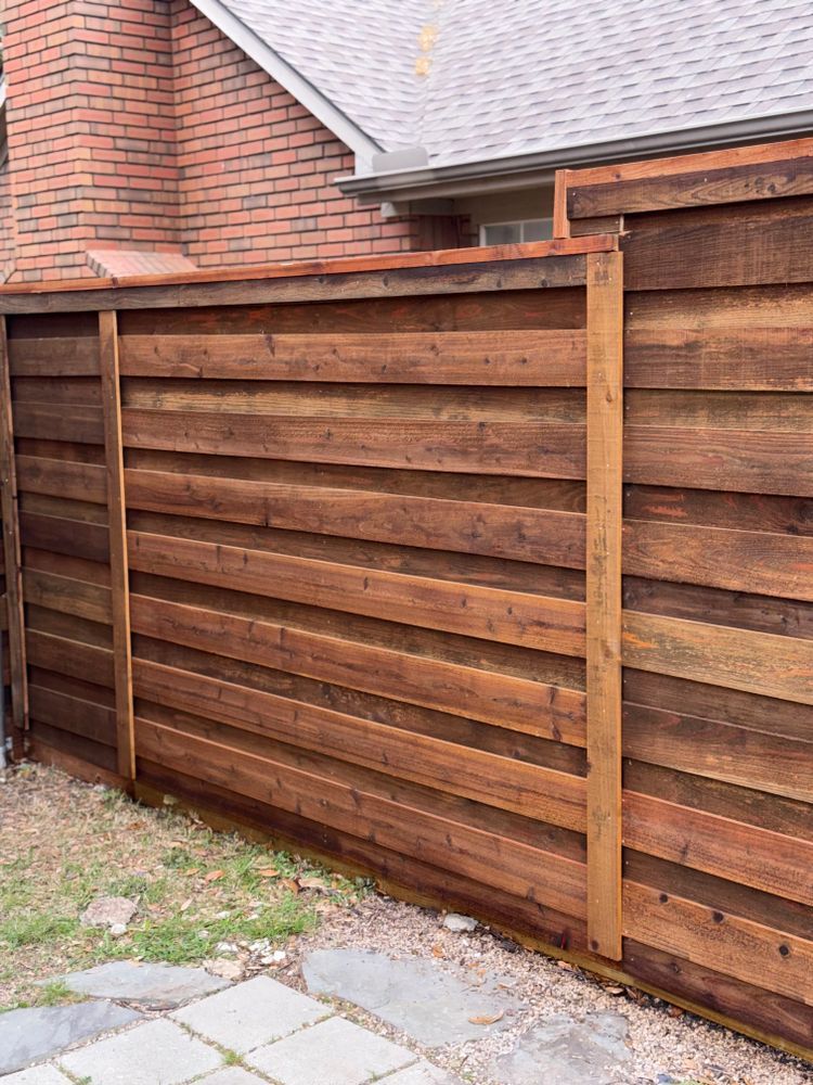Wooden privacy fence beside a brick house, with a stone patio in the foreground.