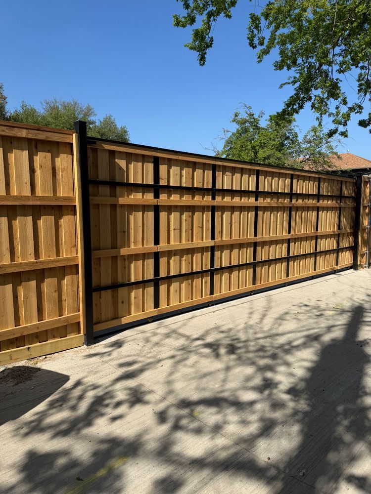 Wooden privacy fence beside a sunlit concrete driveway, with tree shadows and blue sky above.
