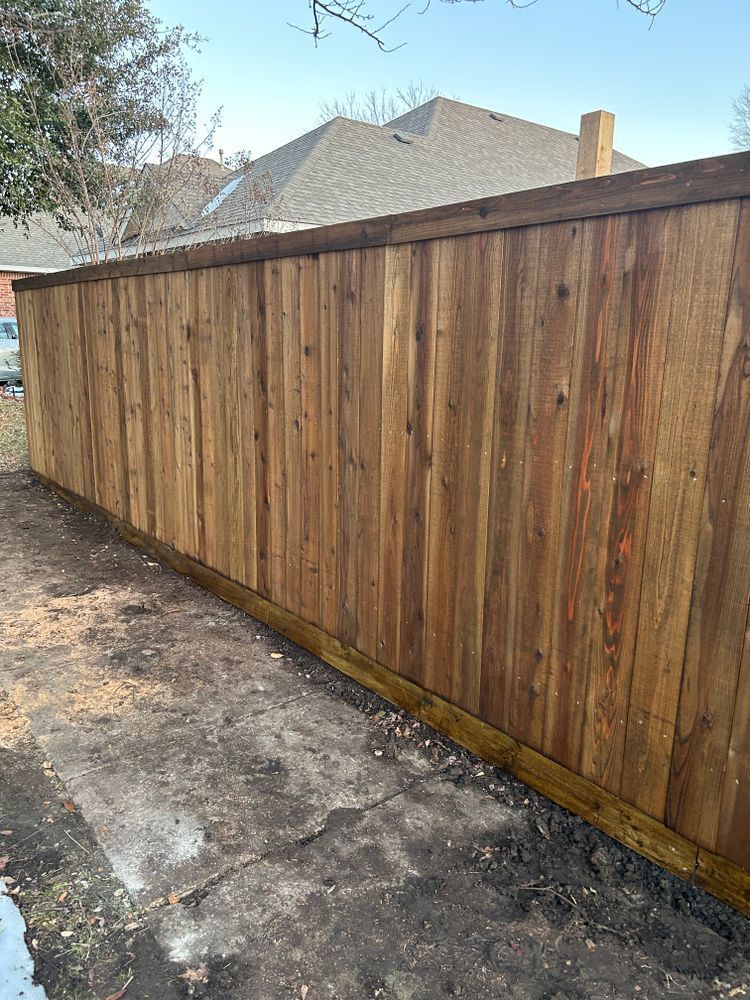 Wooden privacy fence beside a concrete driveway, with houses and trees visible above it.