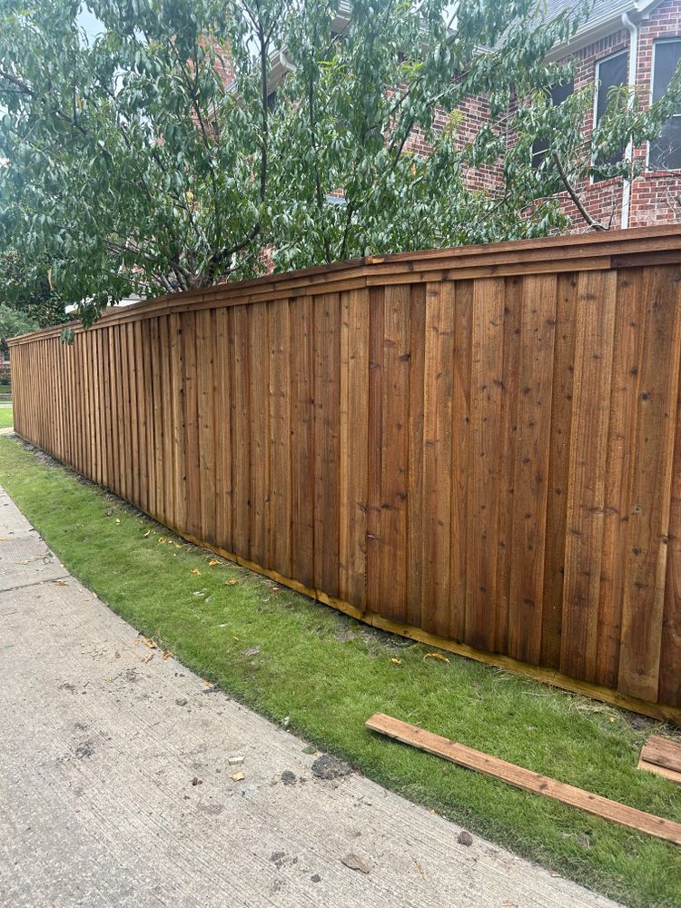Wooden privacy fence along a sidewalk beside a grassy strip, with trees and houses behind it.