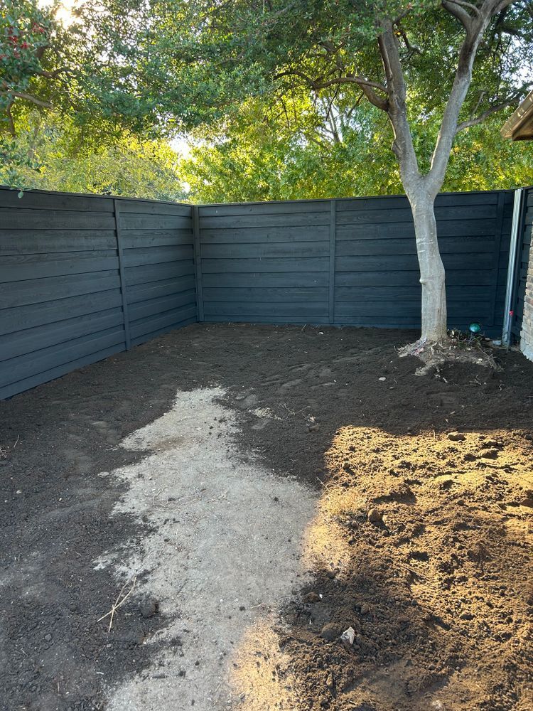 Backyard with dark fence, trees, and bare dirt, including a light gravel path and a small mulched area