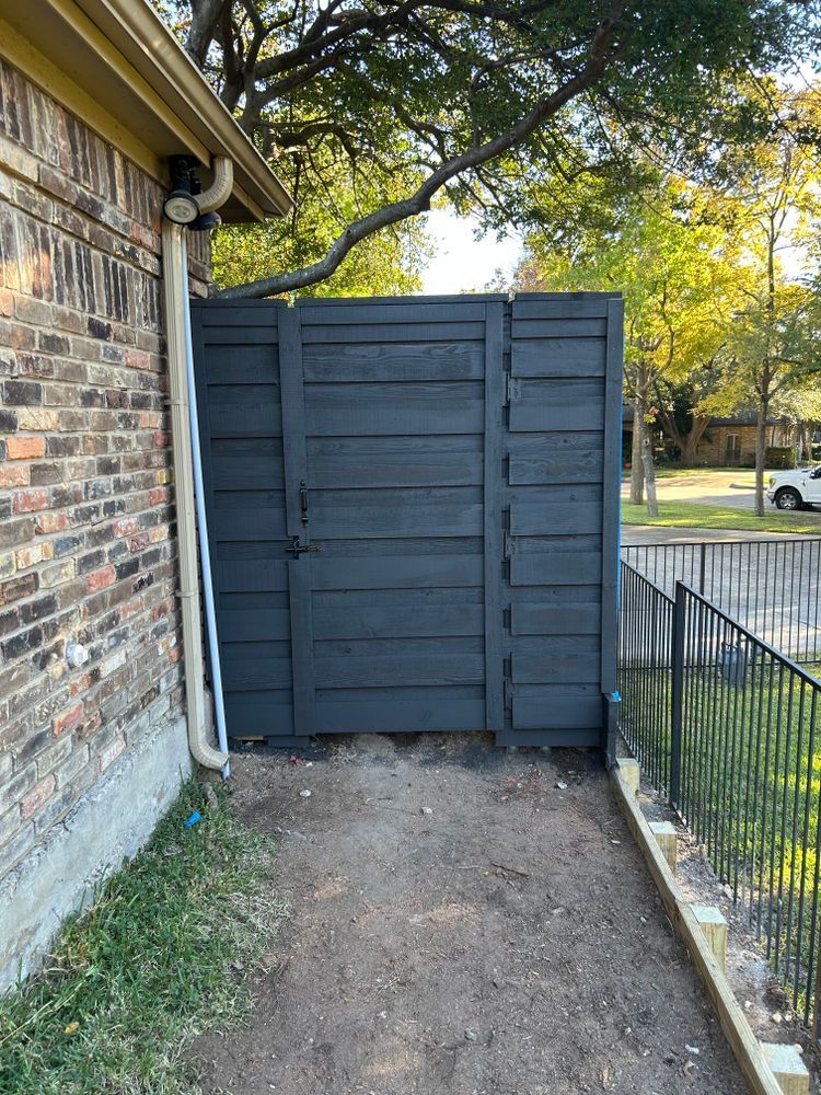 Narrow side yard with dark wooden fence gate beside a brick house and metal railing.