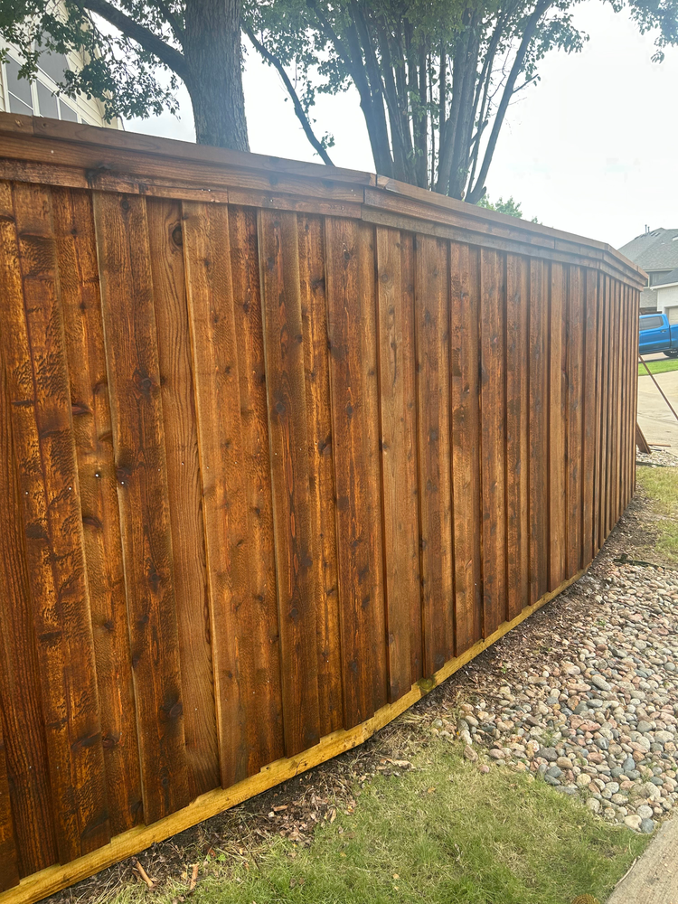 Tall brown wooden fence beside a gravel path and grass, with trees visible above it.