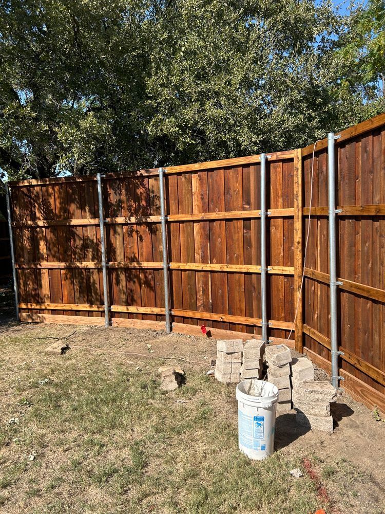 Wooden backyard fence under trees with stacked cinder blocks and paint buckets on dry grass.