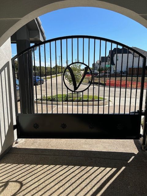 Black metal gate with heart emblem and striped shadows across a driveway under a blue sky