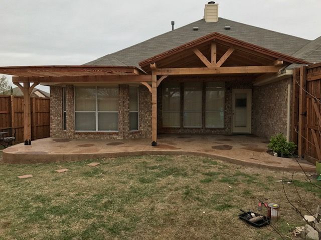 Backyard view of a brick house with a covered patio, wooden beams, and patchy grass.