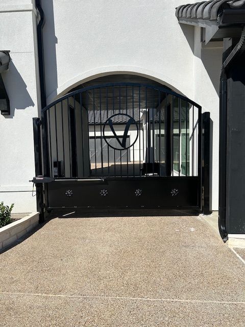 Black wrought-iron gate under an arched entryway, in front of a white house with a textured driveway.