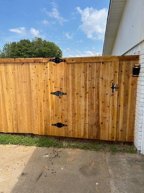 Wooden gate in a driveway beside a white brick house, with trees and blue sky in the background.