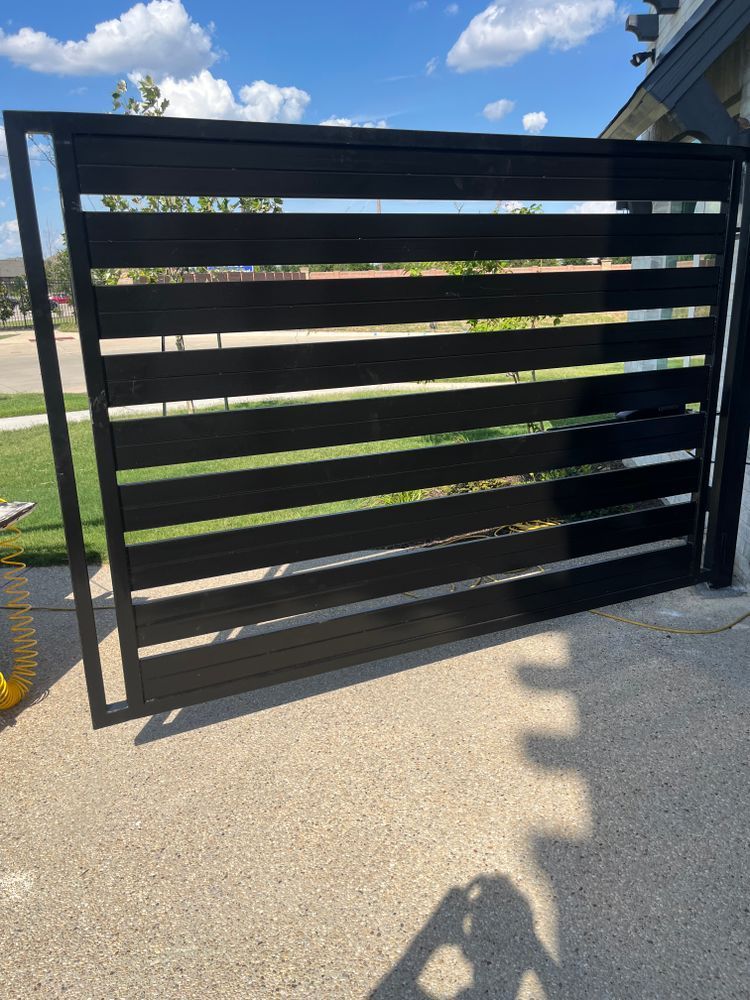Black slatted metal gate with horizontal bars, standing open on a driveway under a sunny blue sky.