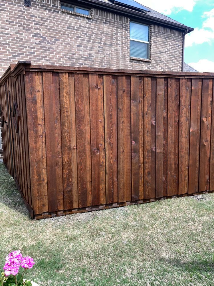 Wooden privacy fence beside a brick house, with grass and a small pink flower in the corner.